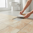 Hands peeling the backing film off a warm beige stone-effect self-adhesive vinyl floor tile and pressing it onto a bathroom floor
