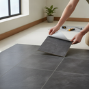 Hands peeling the backing film off a dark charcoal grey stone-effect self-adhesive vinyl floor tile and pressing it onto a hallway floor