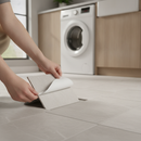 Hands peeling the backing film off a pale light grey stone-effect self-adhesive vinyl floor tile and pressing it onto a utility room floor