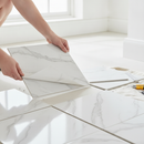 Hands peeling the backing film off a white and grey marble-effect self-adhesive vinyl floor tile and pressing it onto a hallway floor