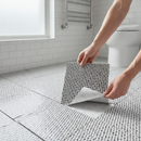 Hands peeling the backing film off a black and white mosaic-patterned self-adhesive vinyl floor tile and pressing it onto a bathroom floor