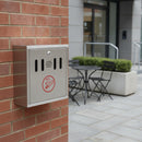 Stainless steel cigarette bin wall mounted on a red brick wall beside a commercial office entrance, with outdoor table and chairs in the designated smoking area behind
