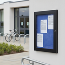 Portrait black lockable notice board fixed to the rendered wall beside a school entrance, blue felt panel visible behind the polycarbonate cover, with bicycle hoops and an access handrail nearby