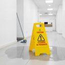 Yellow folding wet floor warning sign standing in an office corridor next to a freshly mopped wet patch on the tiled floor, with a mop leaning against the wall and a reception desk in the background