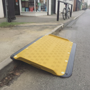 Yellow anti-slip wheelchair kerb access ramp bridging a pavement kerb outside a shop entrance, with a bicycle parked nearby for scale