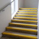 High-visibility yellow PVC stair nosing 50mm x 42mm fitted over the front edge of every concrete step in a public building stairwell, ribbed tread face visible