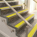 Raked back nosings with bright yellow ribbed inserts on mottled stone steps between stainless handrails