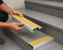 Installer holding a yellow-insert tile-in stair nosing beside one already fitted to a grey tiled step