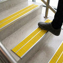 Foot stepping onto concrete stairs fitted with twin yellow anti-slip inserts in aluminium nosings